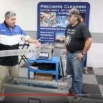 Two men demonstrating industrial parts cleaning equipment